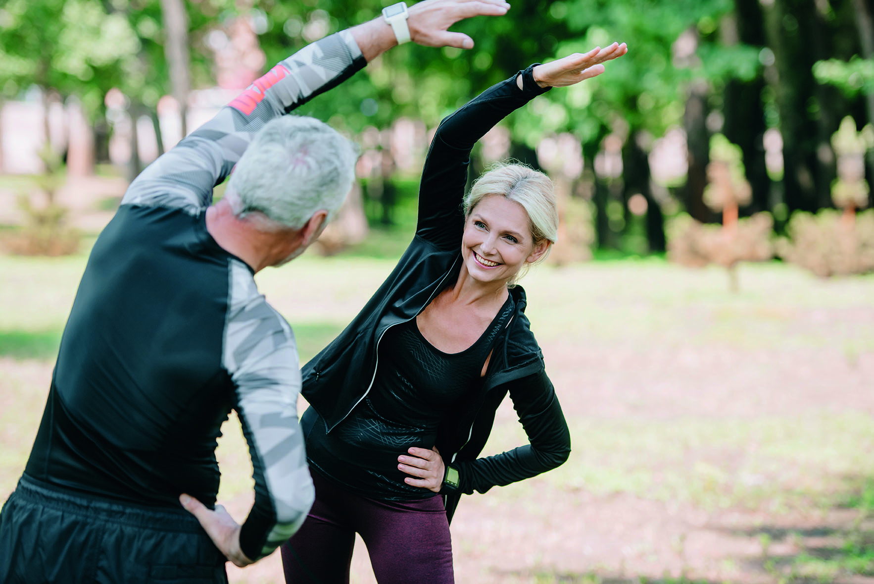 back view of mature sportsman warming up near smiling sportswoman in park