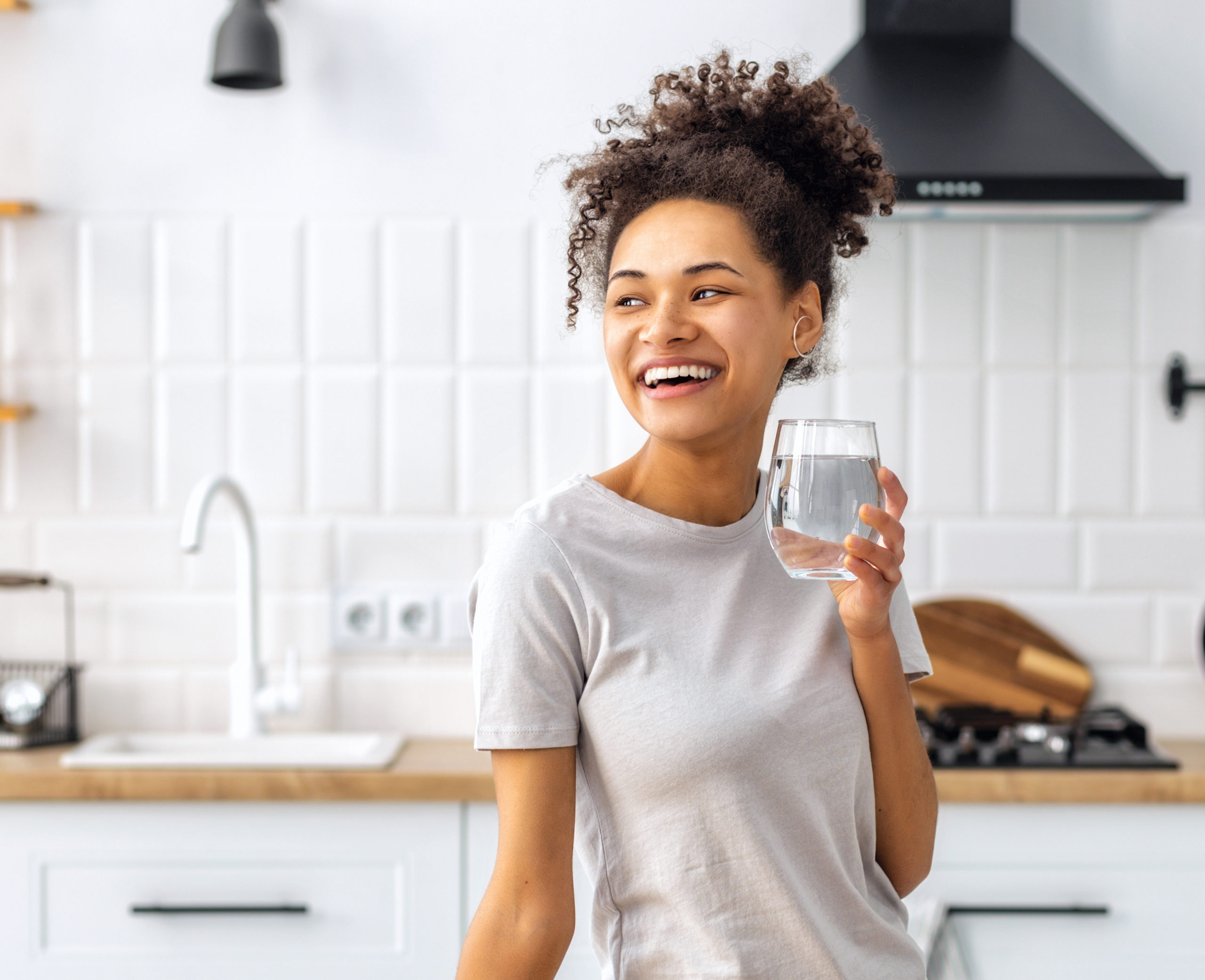 Portrait of beautiful African American young woman drinking water standing in the kitchen at home looking away and smiling, wellness healthy food concept